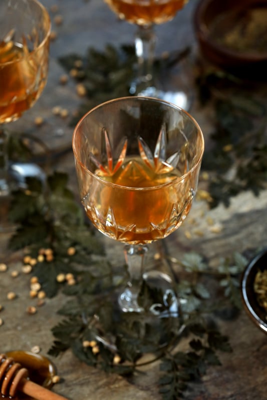 Homemade herbal cordial in a crystal glass surrounded by seeds and honey stick.