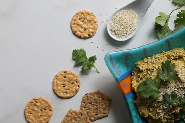 Crackers and spoon of sesame seeds and cilantro leaves on counter next to bowl of carrot veggie dip