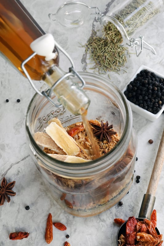 Apple cider vinegar being poured over a variety of herbs to make fire cider at home. Elderberries, whole chilis, astragalus root, star anise, and rosemary.