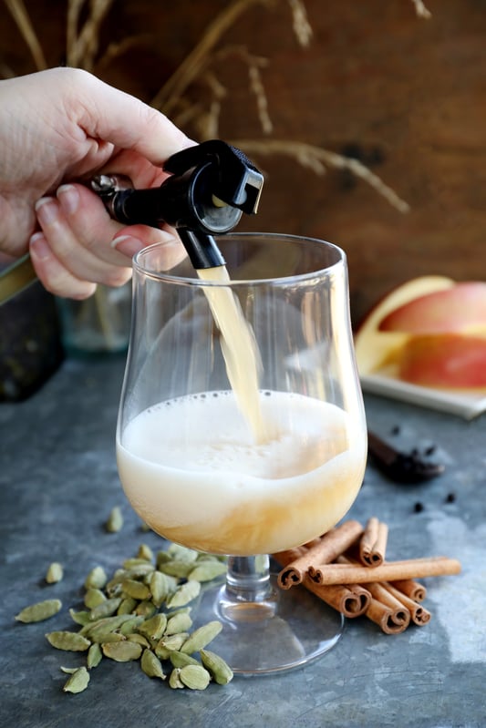 Hand pouring bubbly beverage our of a spout into ornate glass with warm botanicals in background