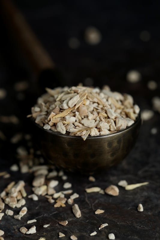 Dried ashwagandha root pieces piled in a polished metal bowl and spilling onto a dark granite surface with a dark background.