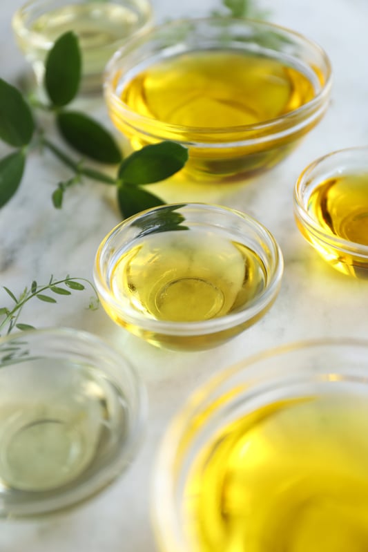 Carrier oils of different shades arranged in clear glass bowls. From opaque white to deep yellow and golden in color, oils are arranged on a white marble counter top with leaves.