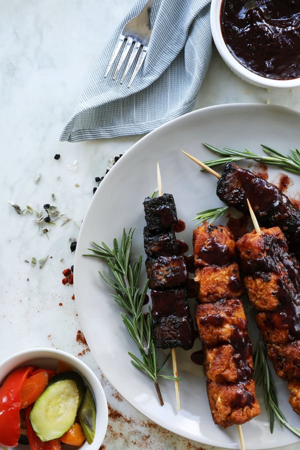 Skewers of tofu are fresh off the grill and smothered in bbq sauce. Served on a plate with fresh herbs and a bowl with fresh vegetables.