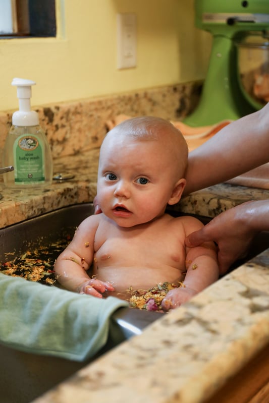 Baby taking bath in sink with natural bath soap behind.