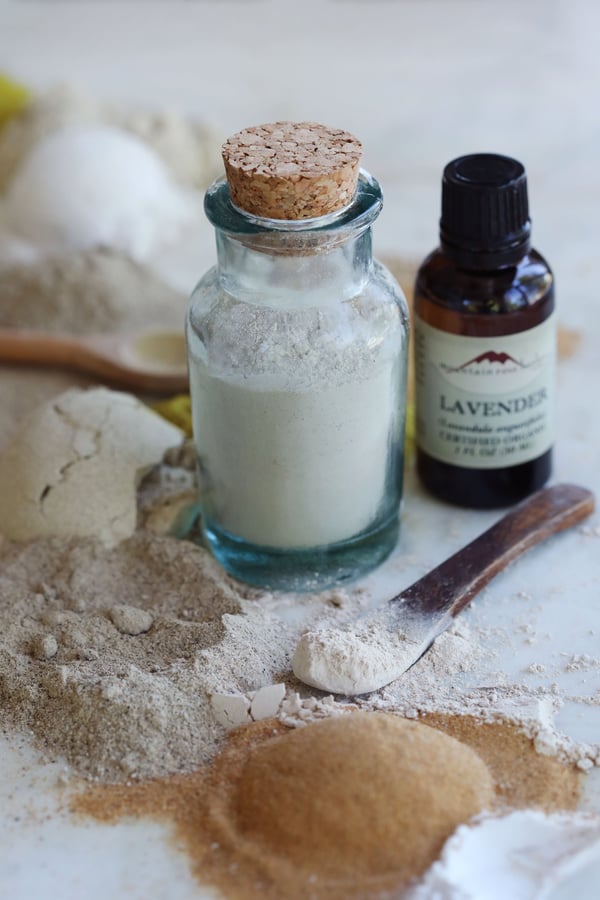 Powdered ingredients are assembled to be made into a homemade, natural baby powder. The finished powder is bottled in an antique glass jar with a cork top, and features a bottle of lavender essential oil. 