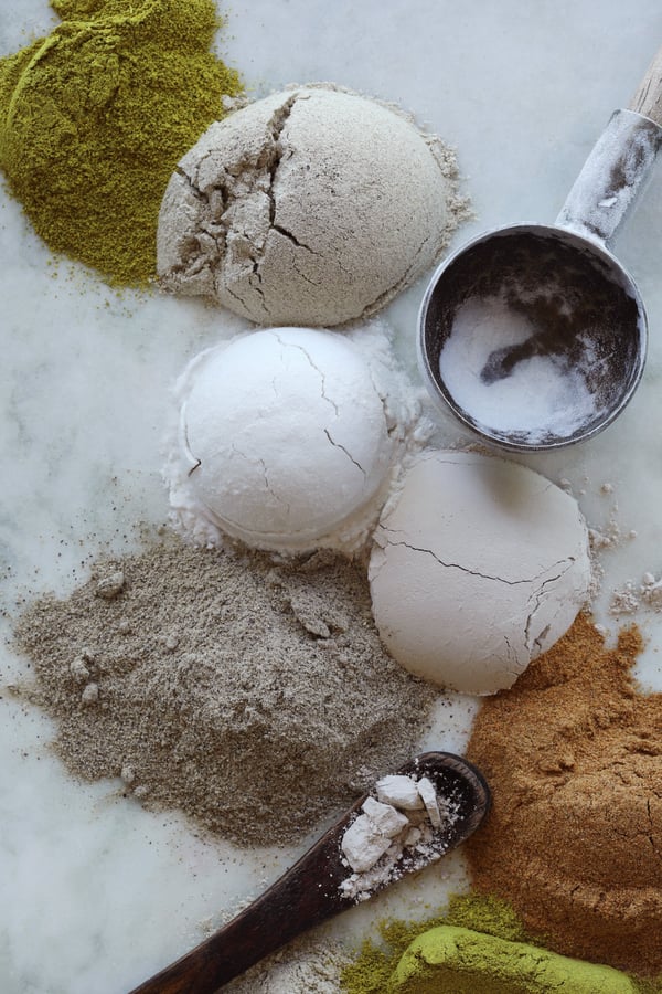 Lumps of cosmetic and herbal powders are assembled with measuring cups and spoons on a white marble counter top. 