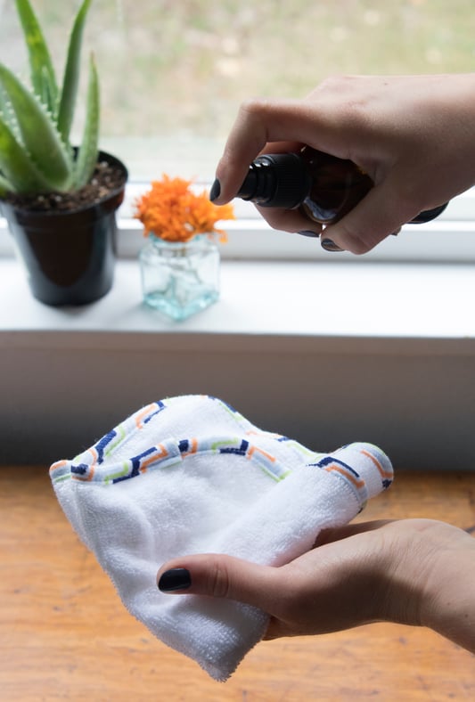 Hand holding baby towel in one hand and baby cleansing spray in the other to make homemade baby wipes