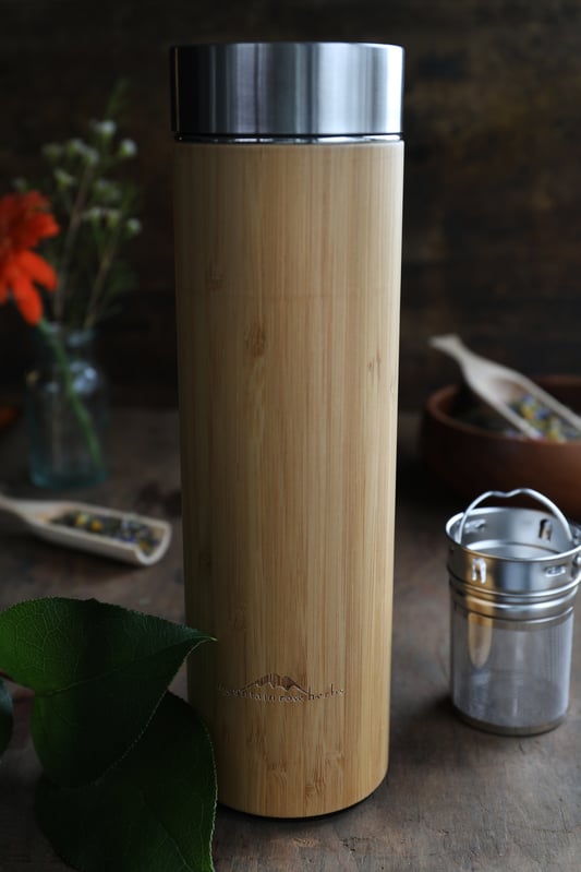 Bamboo and stainless steel tea travel mug with infuser basket on table and fresh plants and herbal teas in background. 