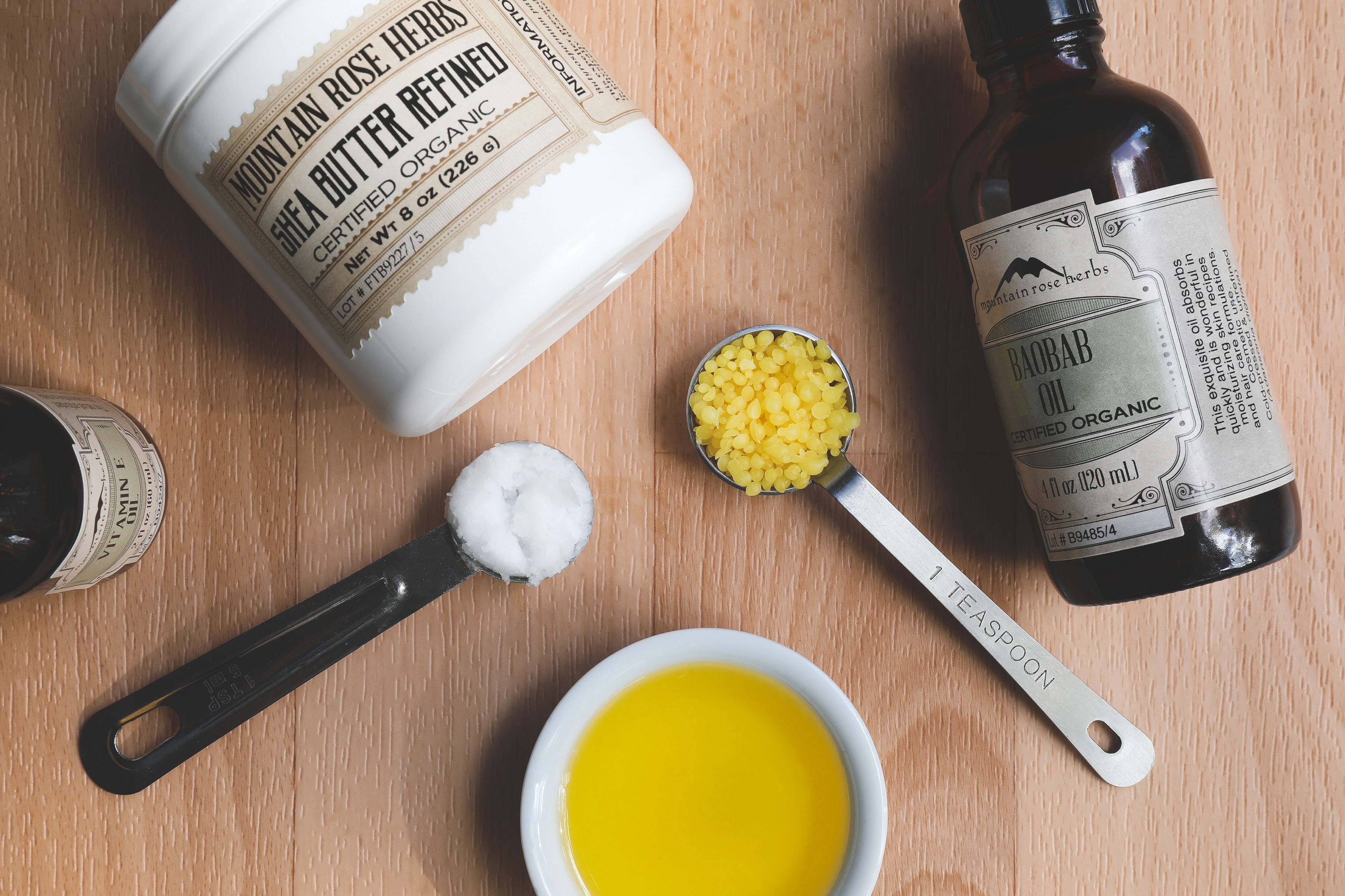 Baobab eye cream ingredients oil in bowl, teaspoon of beeswax pastilles, jar of shea butter, and glass bottles on wood table