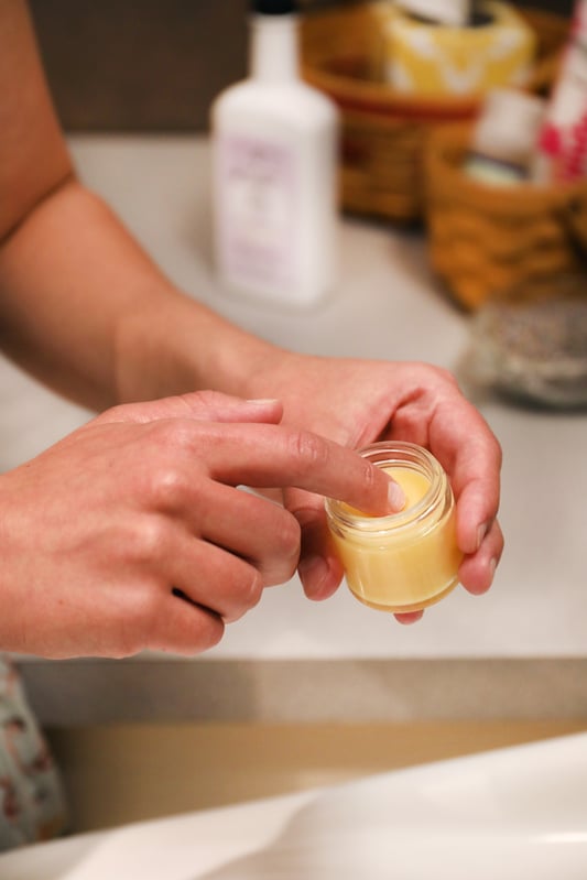 Hands holding open jar of baobab night under eye cream with bathroom toiletries in background