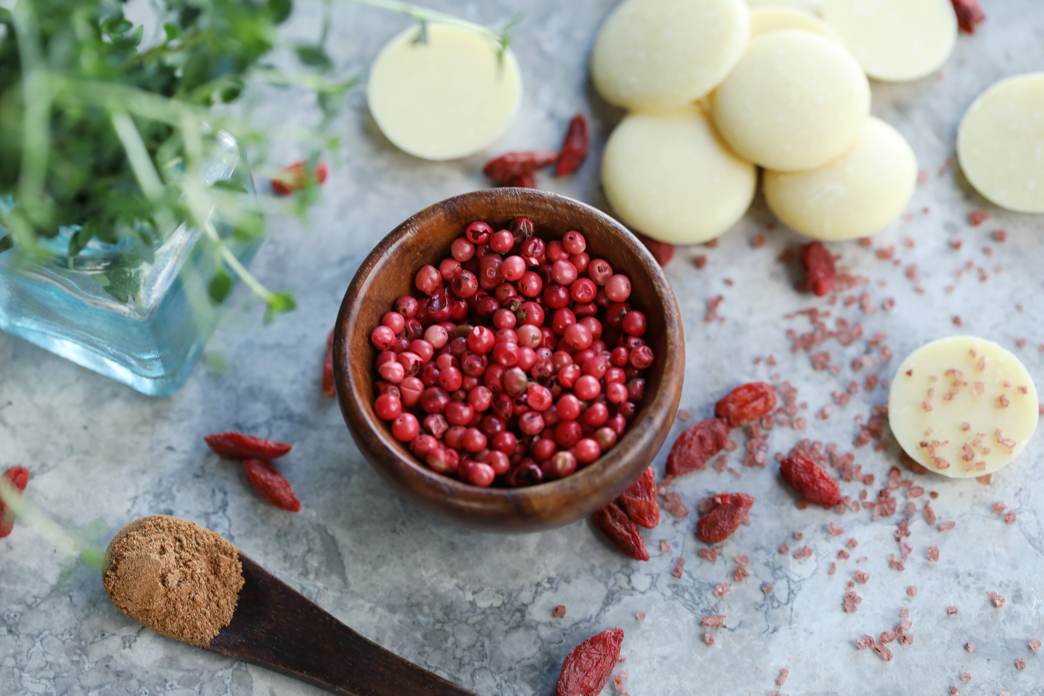 Pink peppercorns in a small wooden bowl with other natural ingredients around with fresh herbs in background. 