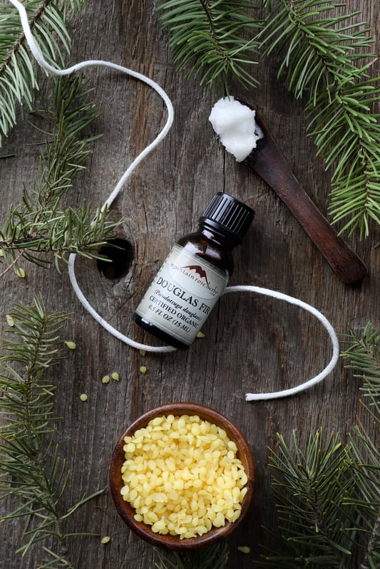 Beeswax pastilles in bowl, bottle of essential oil with candle wick and fir sprigs on wooden table