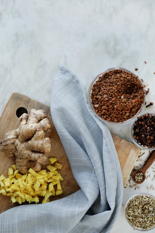 A variety of herbs for wellness support are arranged to be combined in a tincture. Cinnamon, fresh ginger, cloves, and fennel seed on a marble counter top. 