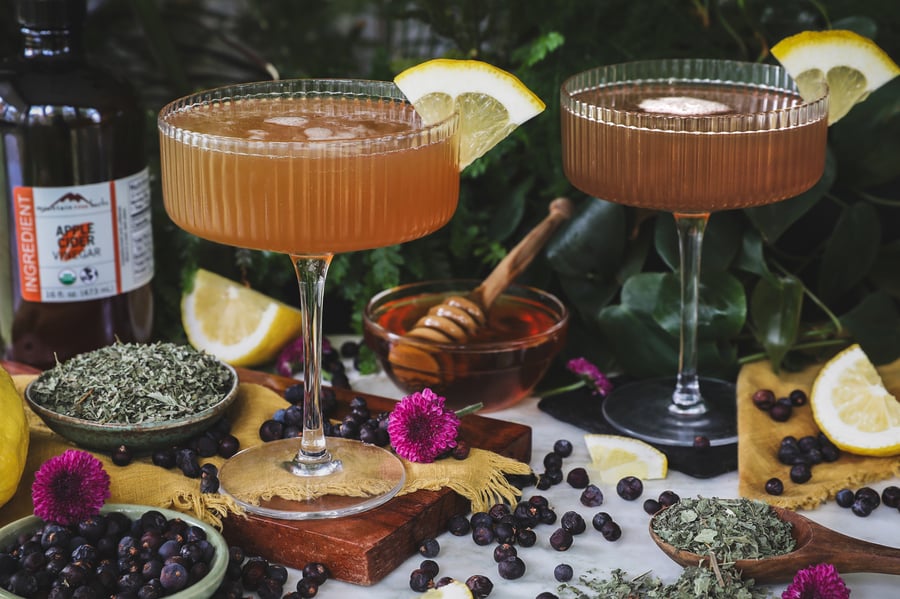 A bees knees mocktail in a stemmed glass surrounded with ingredients and garnished with lemon