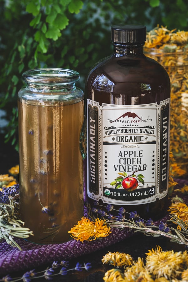 A jar of apple cider vinegar next to a jar with dried herbs and vinegar in it