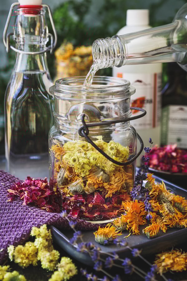 A jar of facial toner herbs with alcohol being poured into it with dried herbs around it