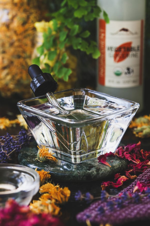 A glass container of vegetable glycerine with a dropper in it and dried herbs nearby