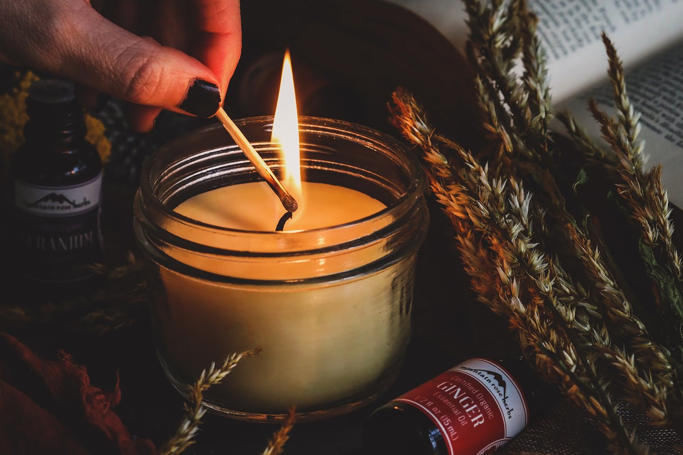 A person lighting a homemade candle in a jar surrounded by botanicals and bottles of essential oils.
