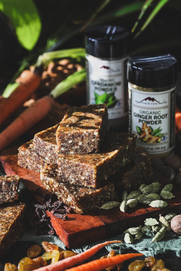 A stack of carrot cake bars sits beside cardamom pods and gold raisins 