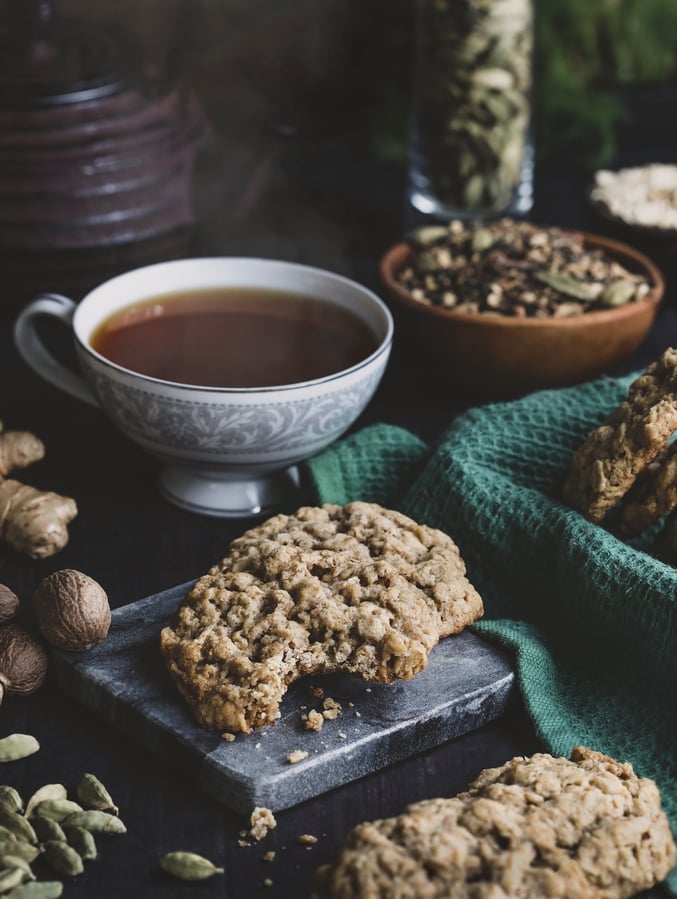 Chai spice oatmeal cookie with a bit out of it, surrounded by cookies, a cup of tea, and whole spices.