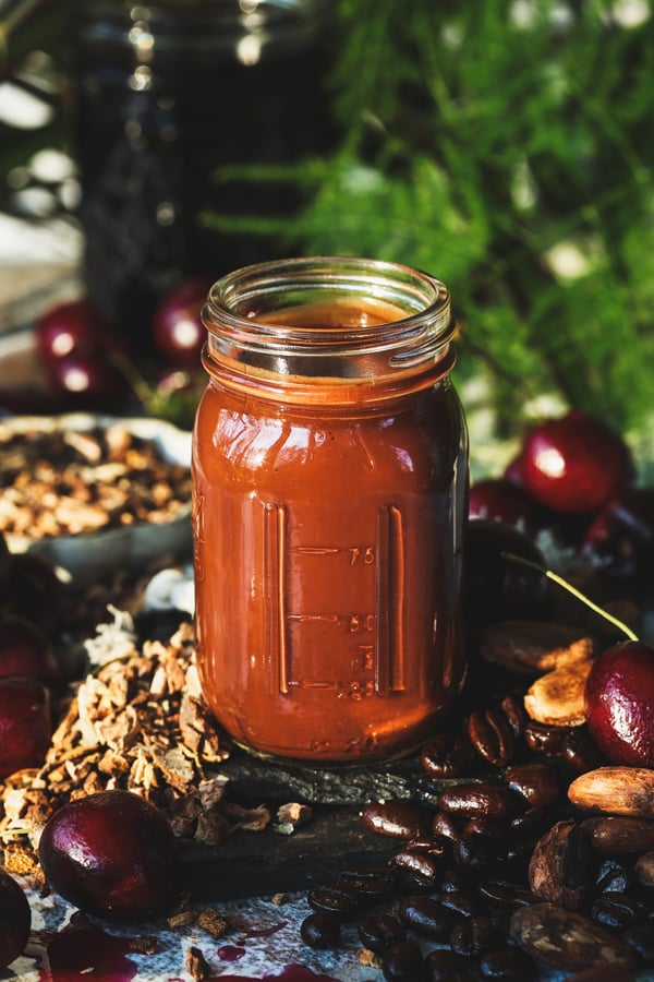 A small jar of cherry chocolate syrup with fresh cherries, cacao bean, wild cherry bark, and coffee beans around it