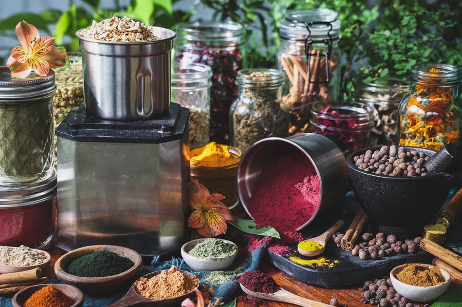 A spice grinder with herbs and herbal powders around it