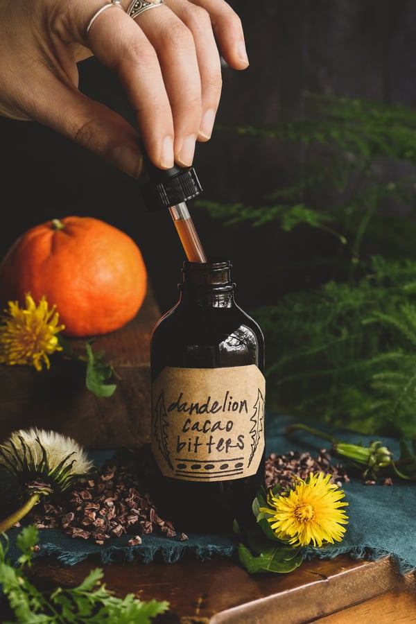 Amber glass bottle with homemade bitters surrounded by fresh dandelion plant and cacao nibs. 