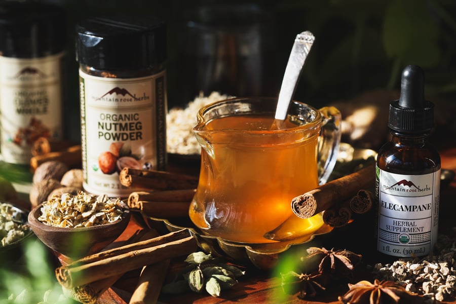 A container of soothing throat syrup with a spoon in it sit with elecampane extract, and chai spices around it