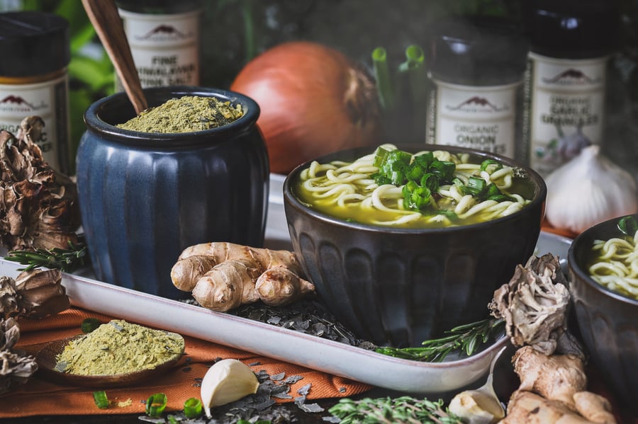 A bowl of ramen beside a container of fire cider bouillon powder with ingredients around it