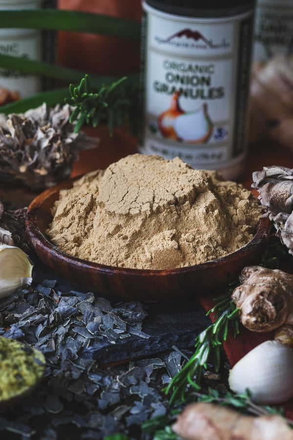 A small bowl of maitake mushroom powder with kombu flakes, ginger, herbs, and garlic around it