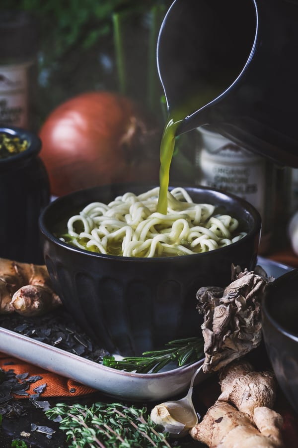 Warm herbal fire cider broth being poured over noodles with ingredients around the bowl