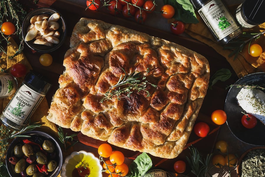 An overhead photo of a full herb focaccia with olives, goat cheese, and olive oil around it