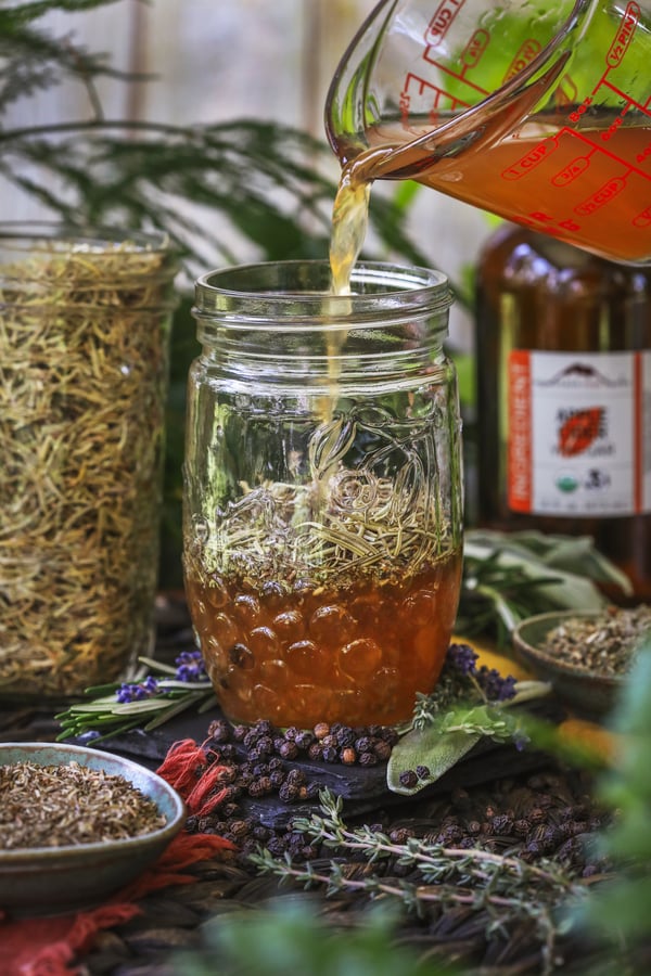 Apple cider vinegar is being poured into a jar of dried four thieves herbs