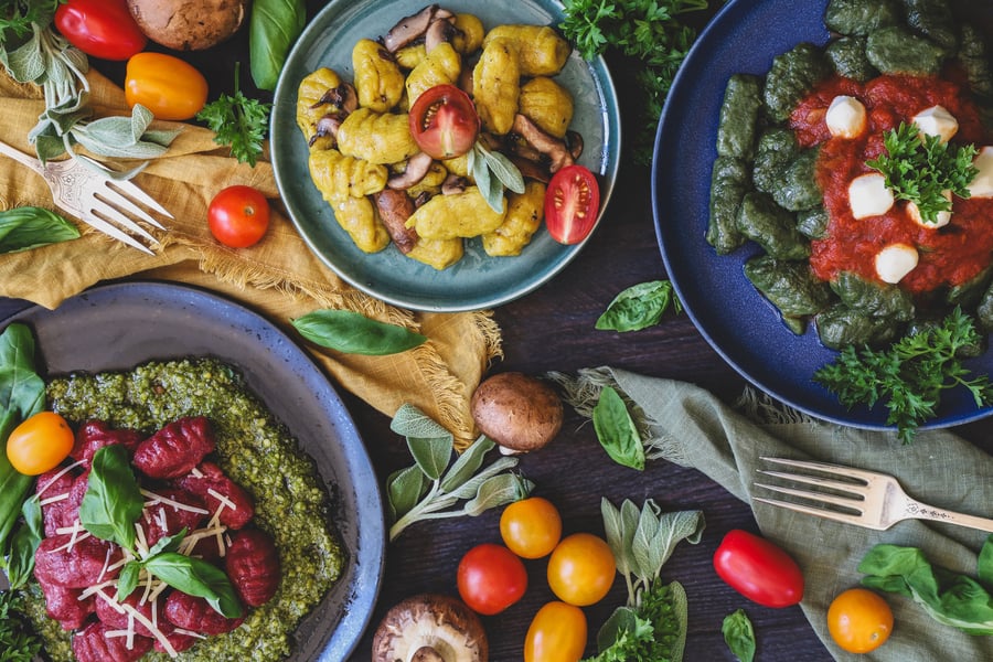 Three different herbal powder gnocchi dishes laid out on a table with fresh parsley, basil, and sage