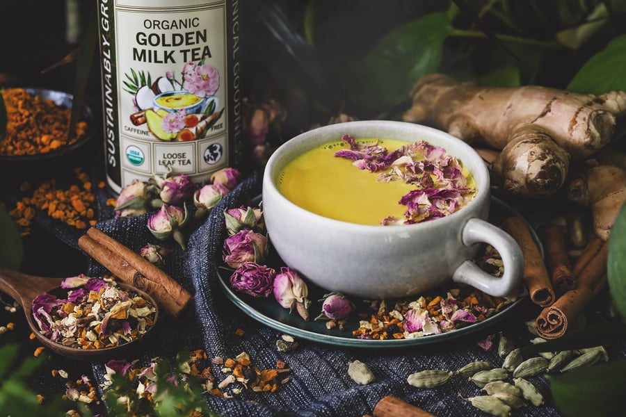 A mug of steamy golden milk with pink rose petals on top surrounded by an array of herbs and spices