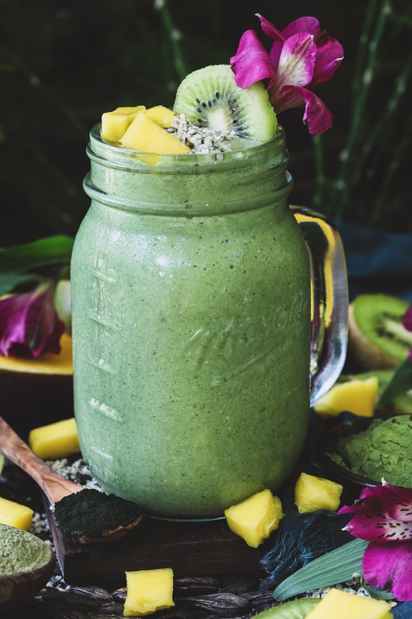 A frosty green smoothie with green powders around it and topped with mango, hemp seeds, and kiwi