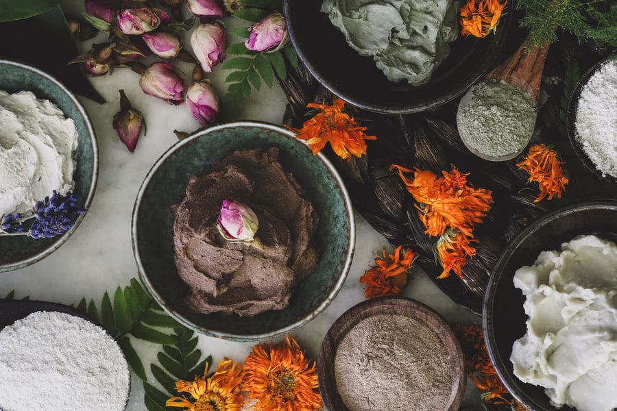 An assortment of dry and hydrated clays in dishes beside skin supporting herbs