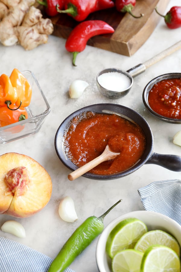 Hot sauce in a bowl surrounded by fresh peaches, colorful peppers, garlic and salt. 