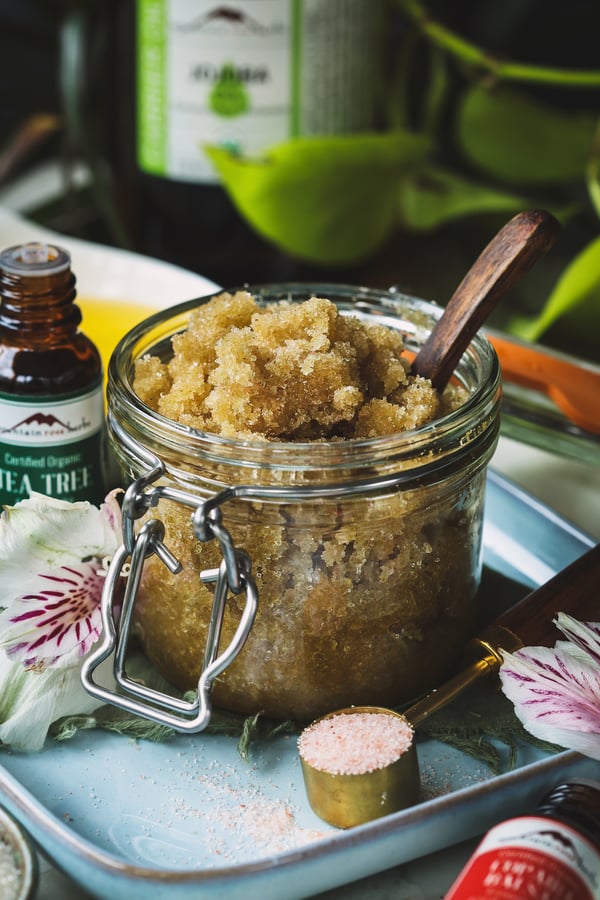 A jar of ingrown hair scrub on a blue tray with flowers, salt and essential oils around it