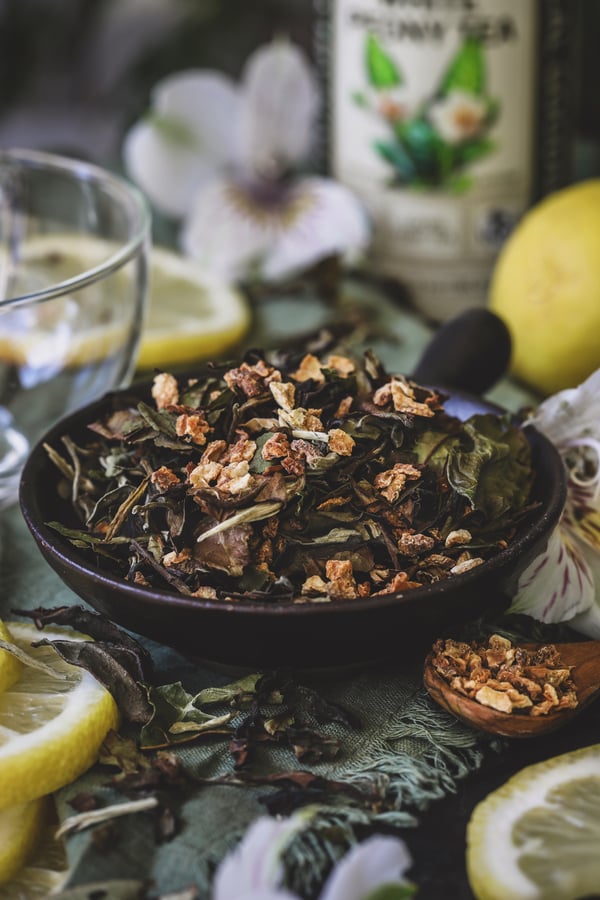 A bowl of white peony mixed with dried lemon peel sits beside an empty tea cup with flowers and lemon slices nearby