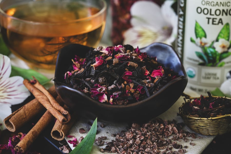 Oolong blended with cacao, cinnamon, and rose petals sites beside a tea infuser, cacao nibs, and cinnamon sticks