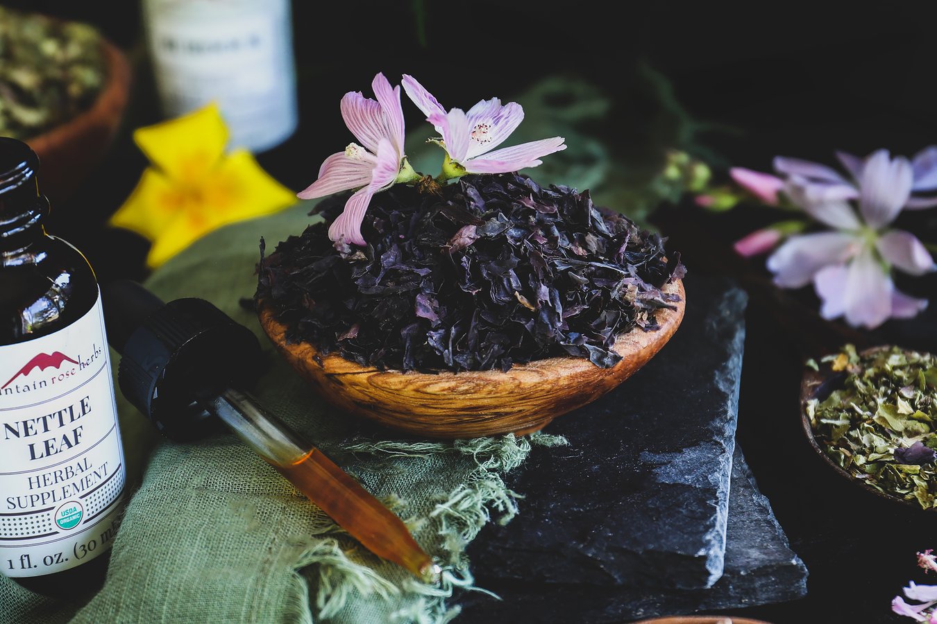 Nori seaweed flakes in a small wooken bowl surrounded by herbs, flowers, and tincture bottle. 