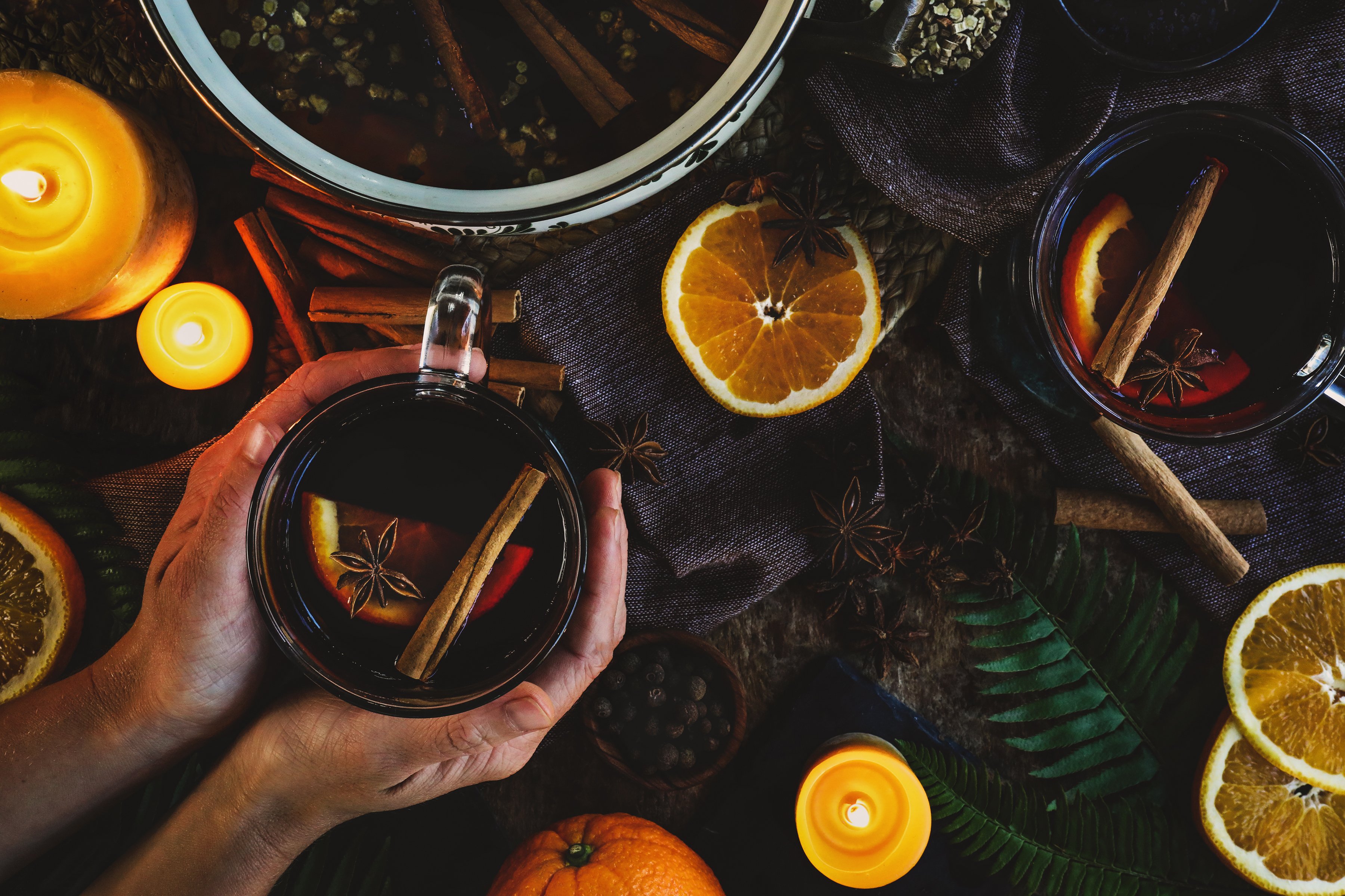 Hands are wrapped around a warm mug of mulled cider with lit candles and orange slices scattered on the table.