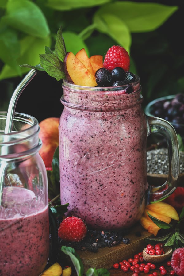 A pink berry smoothie with bilberries and pink peppercorns around it, topped with peach slices and berries