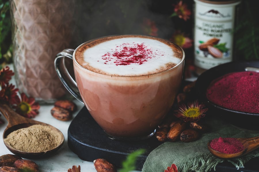 A warm red velvet latte sits among beet root powder, cacao beans, and dandelion root powder