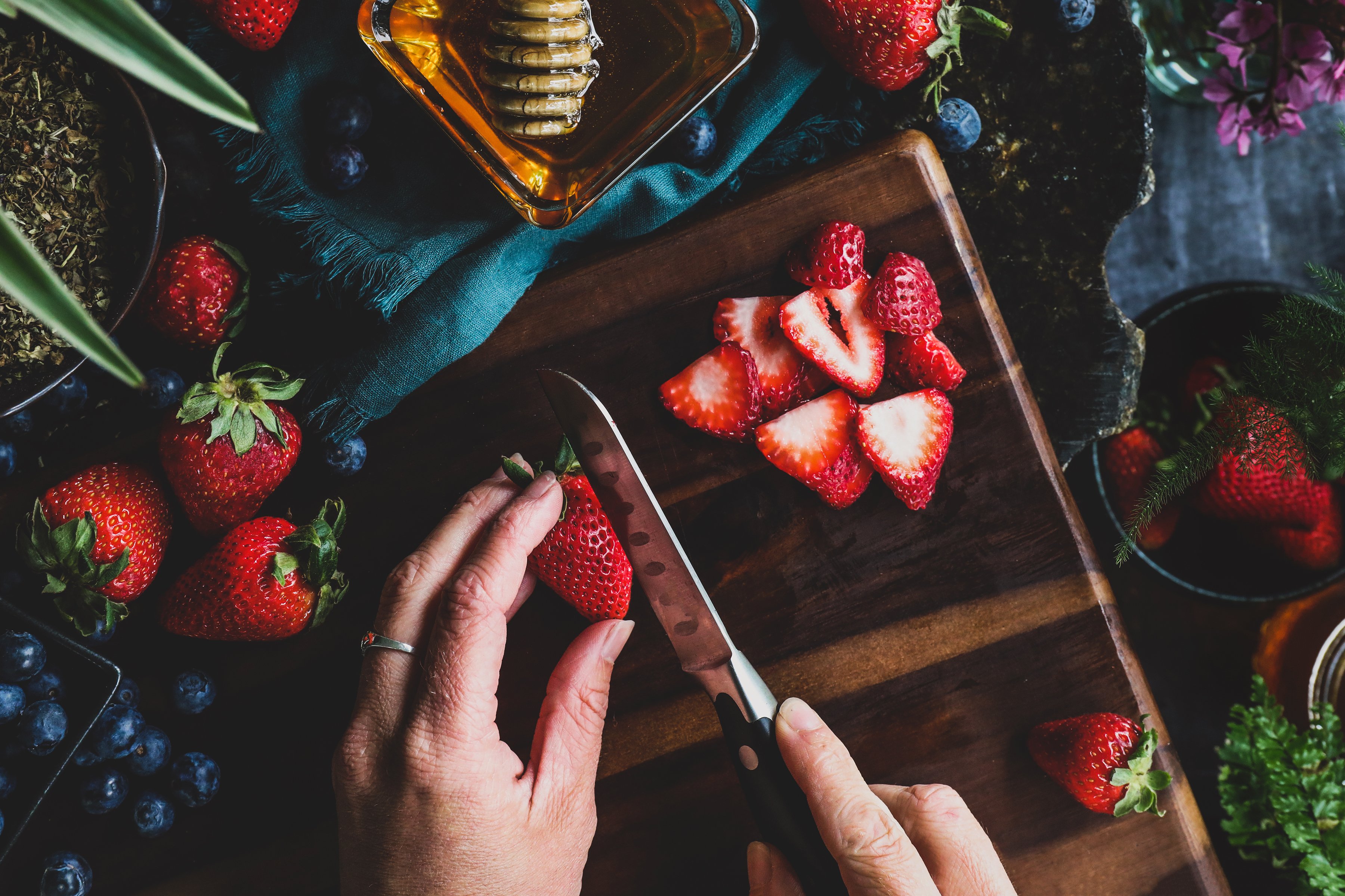 Hands cutting fresh strawberries, surrounded by blueberries and other fresh ingredients. 