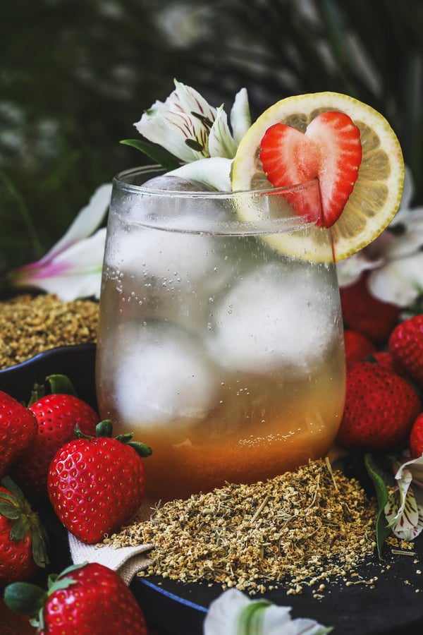 A shrub mocktail sits surrounded with elderflowers and fresh strawberries