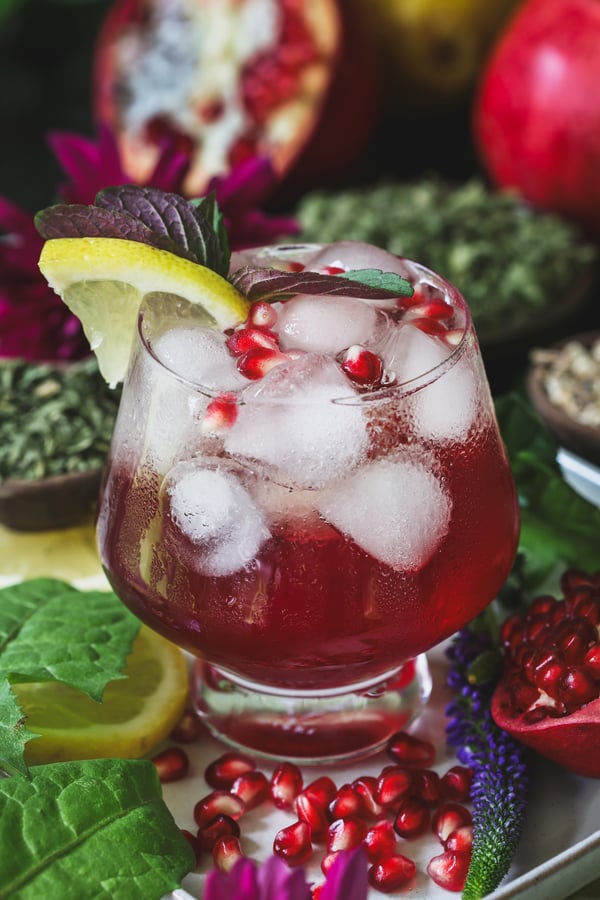 A close up of a spring rejuvenation mocktail with dandelion leaves and pomegranate kernels around it