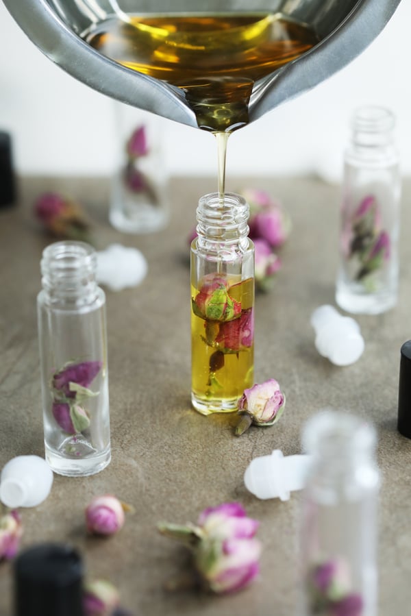 Golden liquid lip gloss pouring from a metal double boiler into a bottle with rosebuds inside, roll-tops on the table ready to go on the bottles.