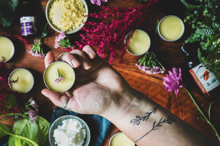 A hand hold a tin of homemade tattoo balm above a table that is strewn with ingredients and flowers